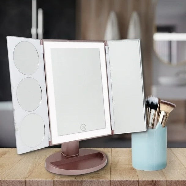 Cosmetic vanity mirror with a folding three-panel design, lighted center panel, and a rose-gold base on a wooden surface with a blue makeup brush holder.