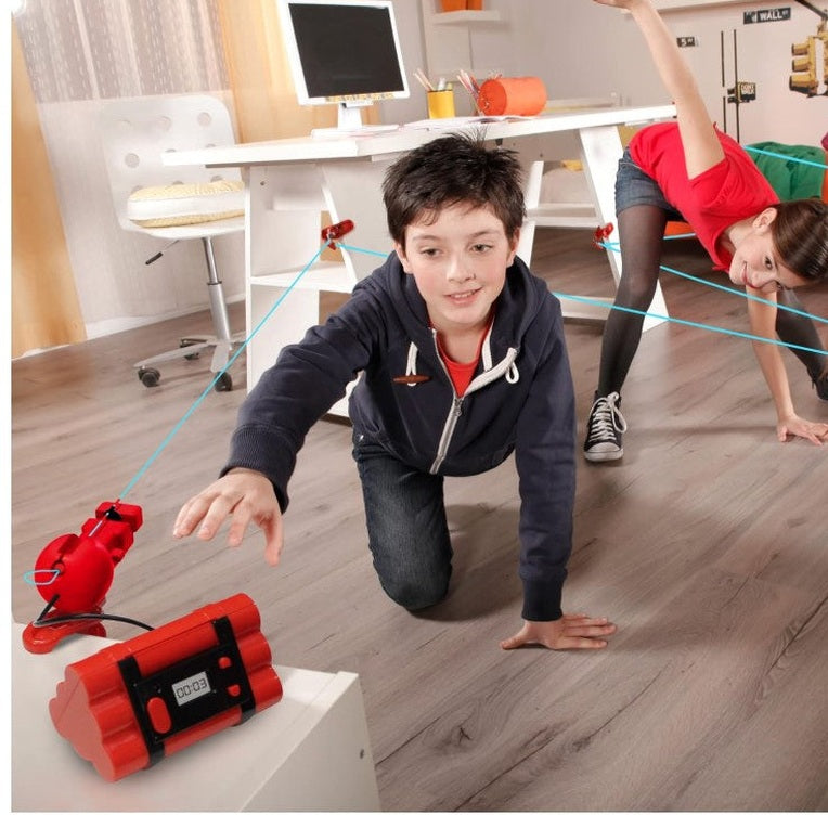Two children crawling on a wood floor toward a red toy bomb with a timer, navigating a blue laser-beam maze set up in a room with white furniture.