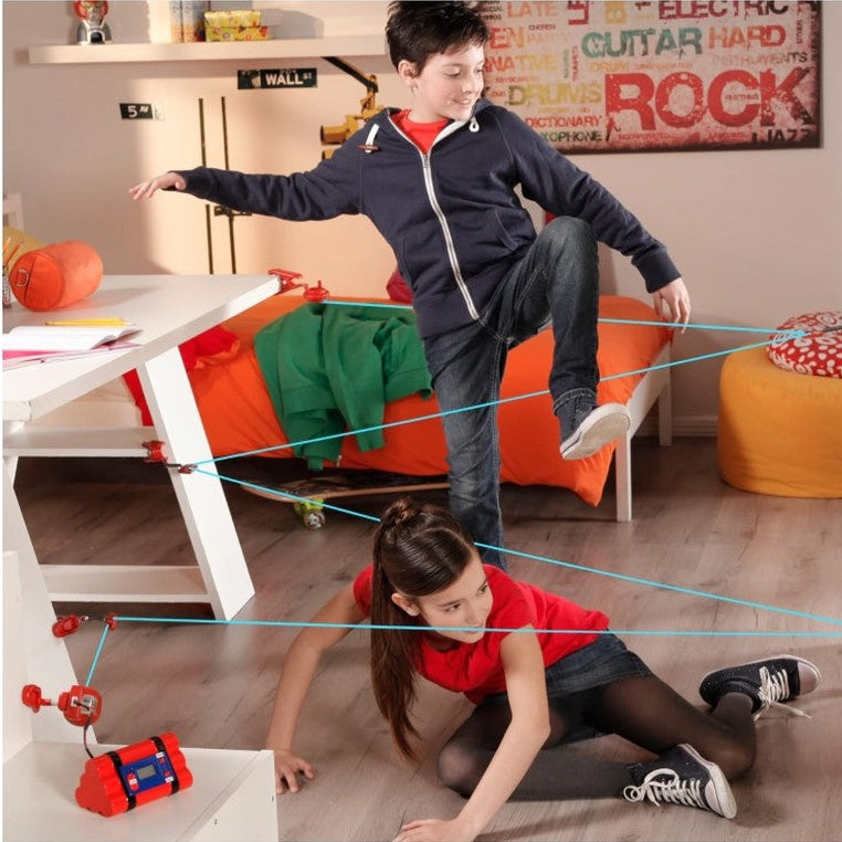 A girl crawling under blue laser beams, with a boy stepping over them in a bedroom with a desk, bed, and wall decorations.