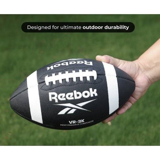 A black Reebok football with white stripes being held by a hand in front of green grass.