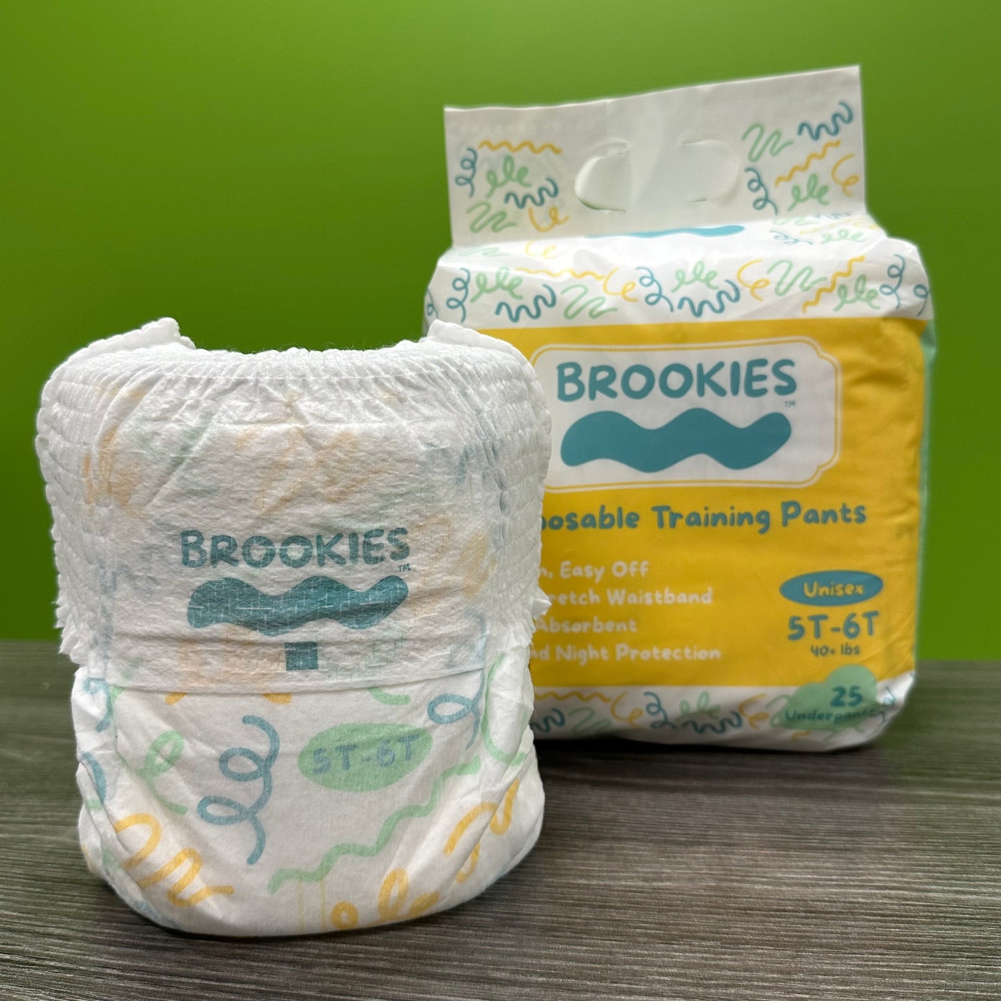 A package of Brookies training pants sits behind a single training pant on a wooden surface in front of a green background.