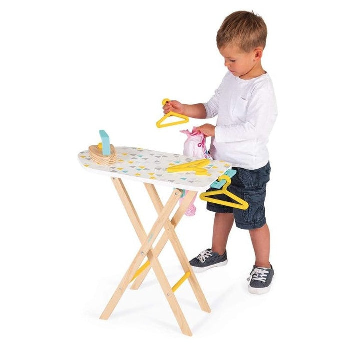 A young boy plays with a toy ironing board, wooden hangers, and a small bag of clothes. He wears a white long-sleeved shirt, denim shorts, and grey sneakers.