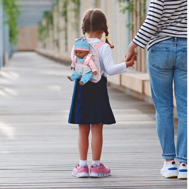 A young girl wearing a dark skirt, a white long-sleeved shirt, and pink sneakers holds hands with an adult in jeans and a striped shirt. The girl has a doll in a carrier backpack. They are on a wooden boardwalk or path.