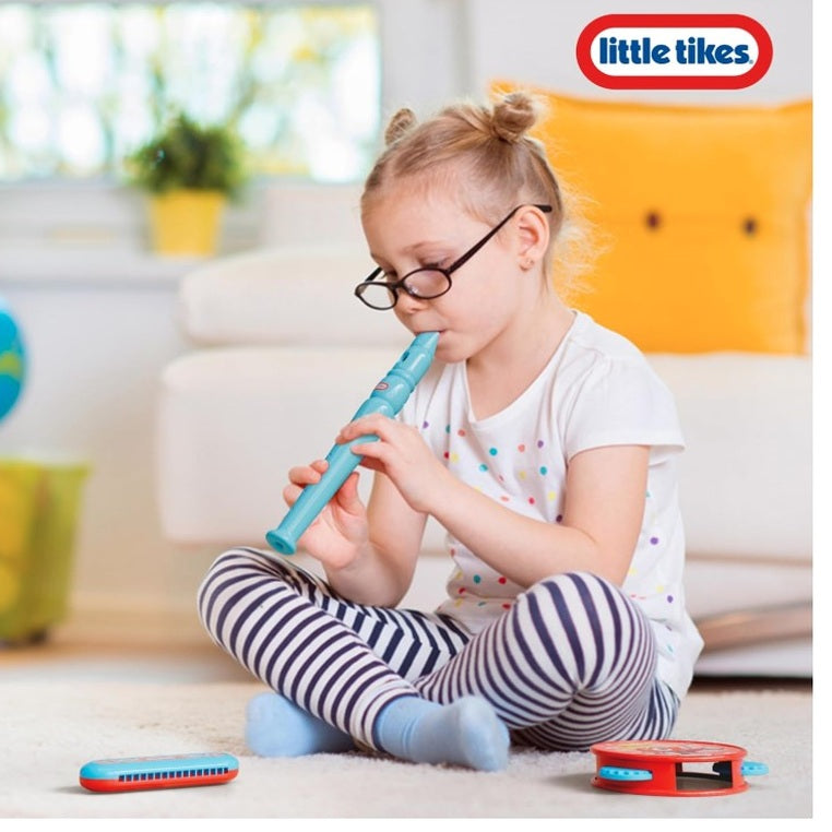 Young girl with glasses playing a blue recorder, wearing a spotted white shirt and striped leggings, sitting on a carpet with a harmonica and tambourine nearby, with a "Little Tikes" logo visible.