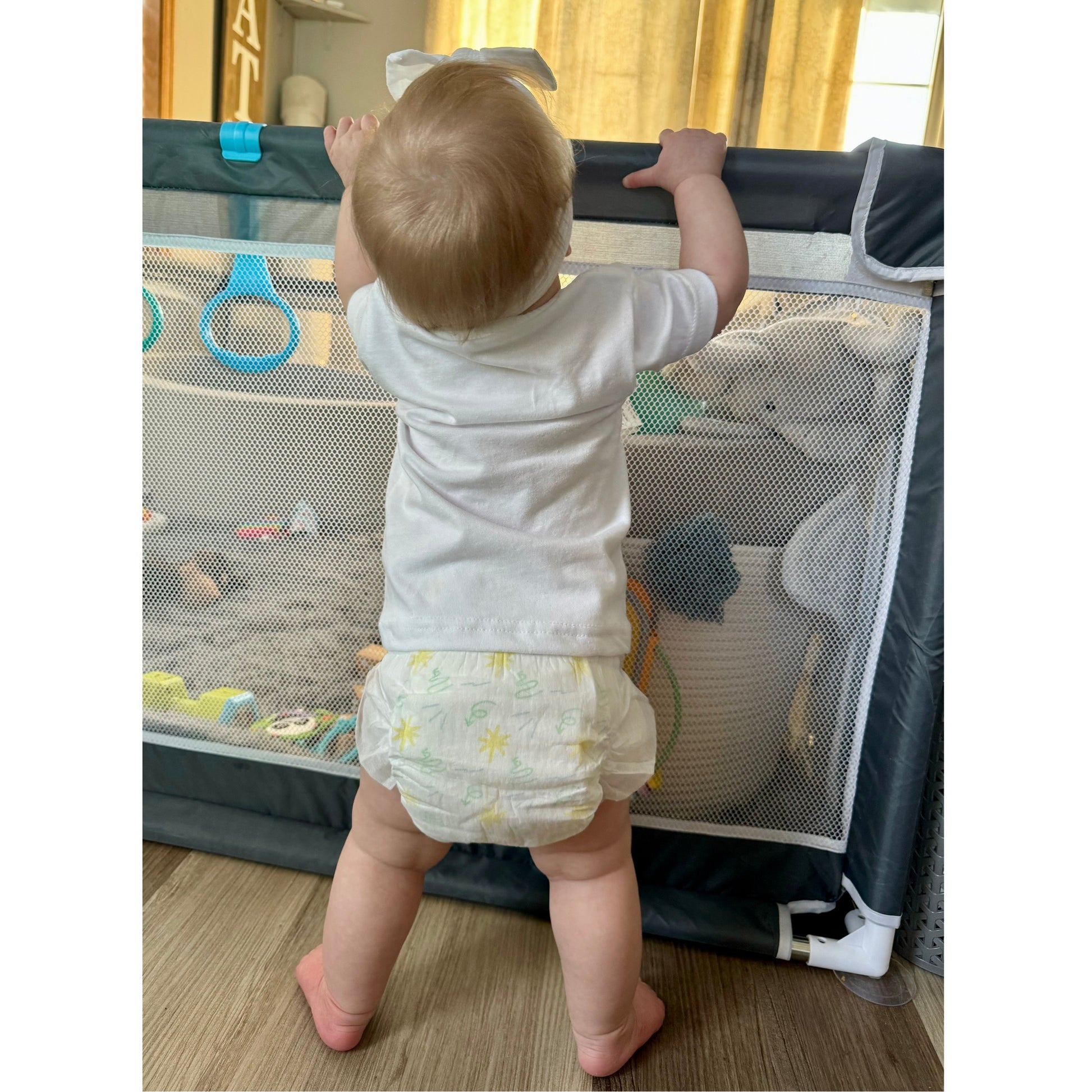 Baby standing in diaper and white t-shirt at a gray playpen with mesh walls.