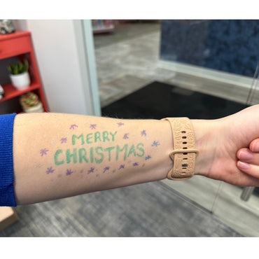 Arm with "Merry Christmas" written in green, surrounded by purple and silver snowflakes, wearing a beige floral-patterned watchband.