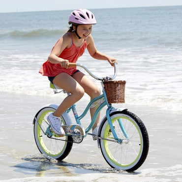 A young girl wearing a pink helmet and red shirt is riding a light blue bicycle with a wicker basket on the beach.