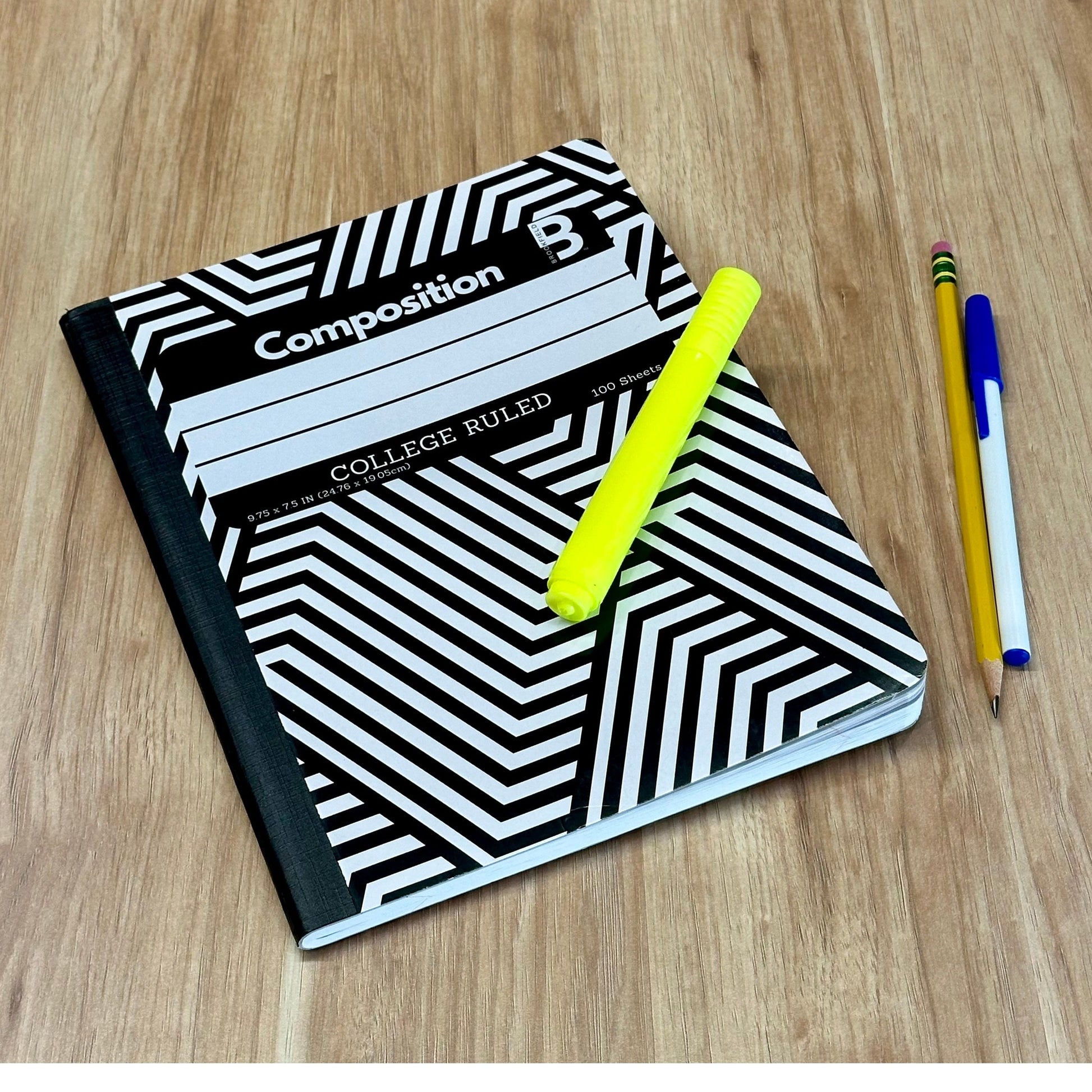 Composition notebook with a black-and-white geometric cover, a yellow highlighter, a pencil, and a blue ballpoint pen on a wooden surface.