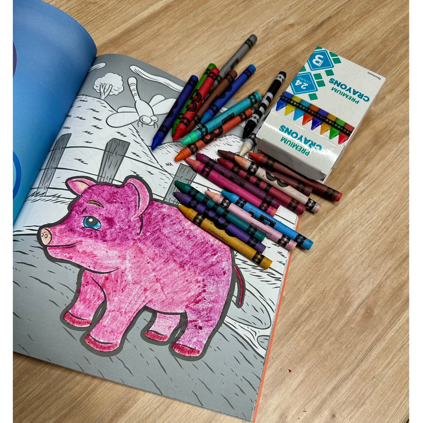 Close-up of a coloring book with a partially colored pink pig, surrounded by scattered crayons and a box of "Premium Crayons."