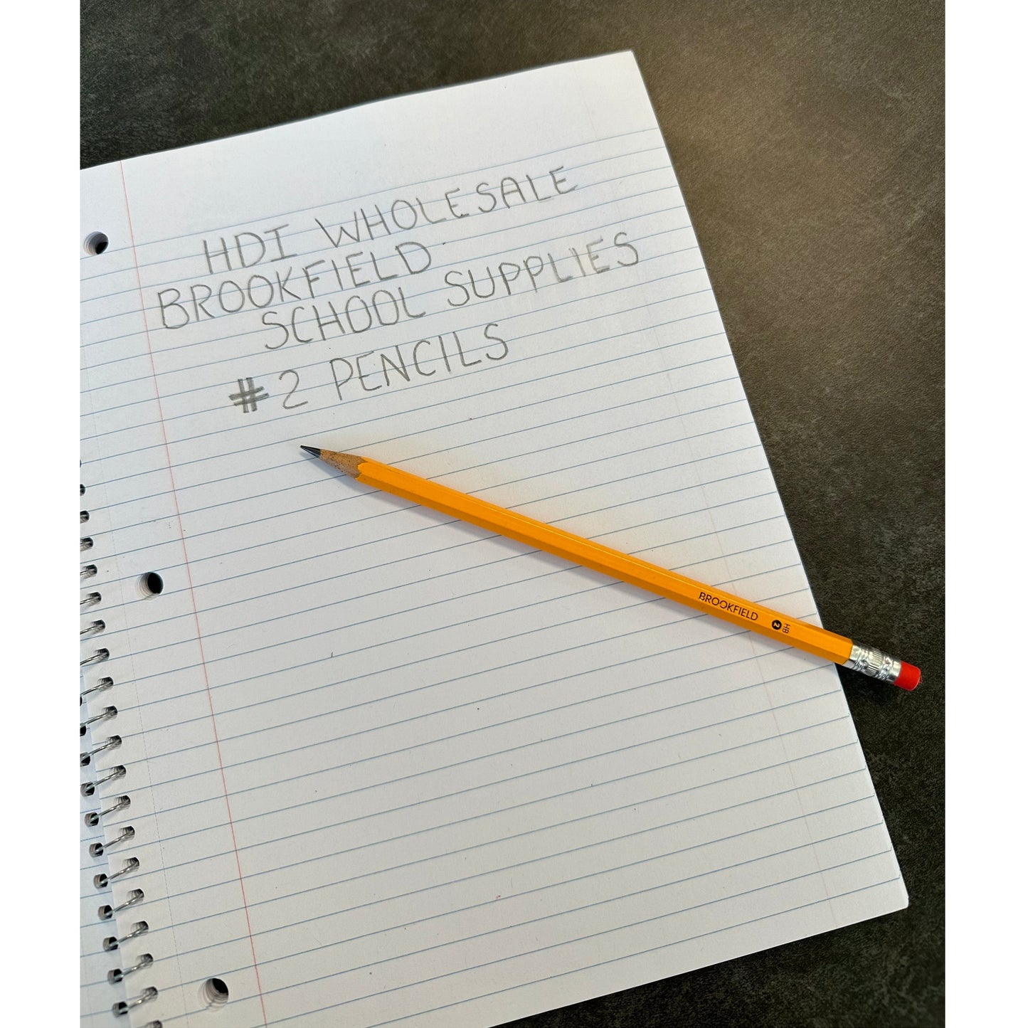 A yellow pencil lies on a notebook with handwritten text that says HDI Wholesale, Brookfield, School Supplies, and #2 Pencils.