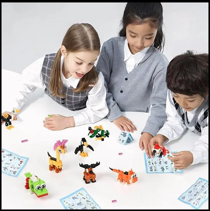 Three young children playing with colorful building block toys on a white table. The children are wearing casual clothes, with two in sweaters and one in a vest. There are various animal-shaped figures made from the blocks spread out around them.
