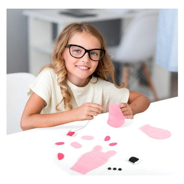 Girl wearing glasses sewing pink felt craft kit at white table.