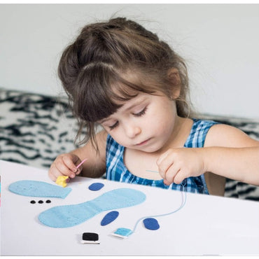 A young girl is intently sewing with blue felt shapes, thread, a needle, and a yellow needle threader on a white table. She is wearing a blue-and-white plaid dress.