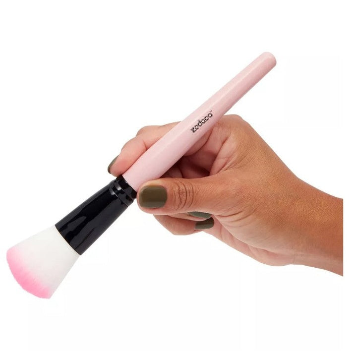A hand holds a makeup brush with a pink handle and pink-tipped white bristles.
