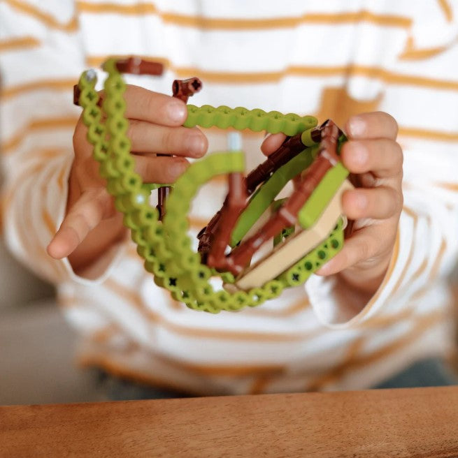 A child holding a wooden and green plastic toy. The child wears a white and yellow striped shirt.