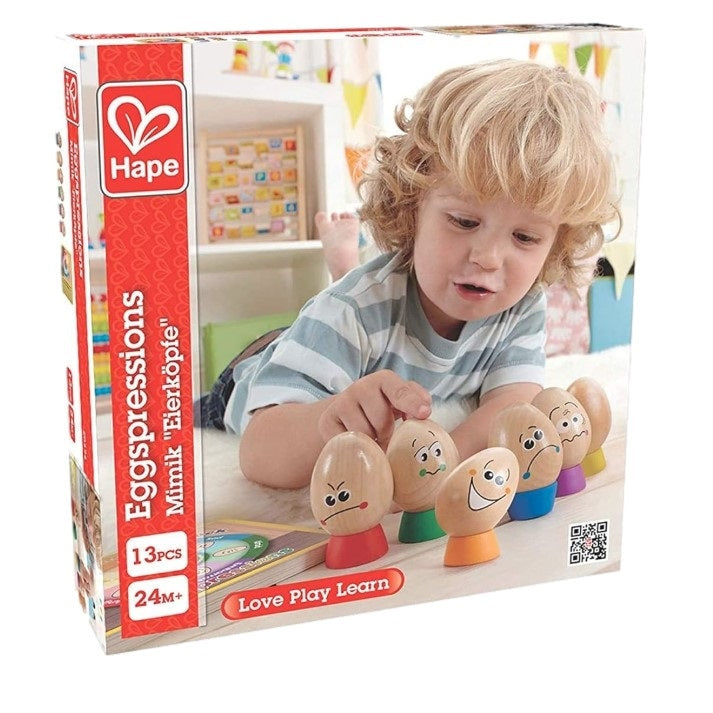Box of Hape Eggspressions toy, with blond toddler in striped shirt playing with the toy eggs on a white surface.