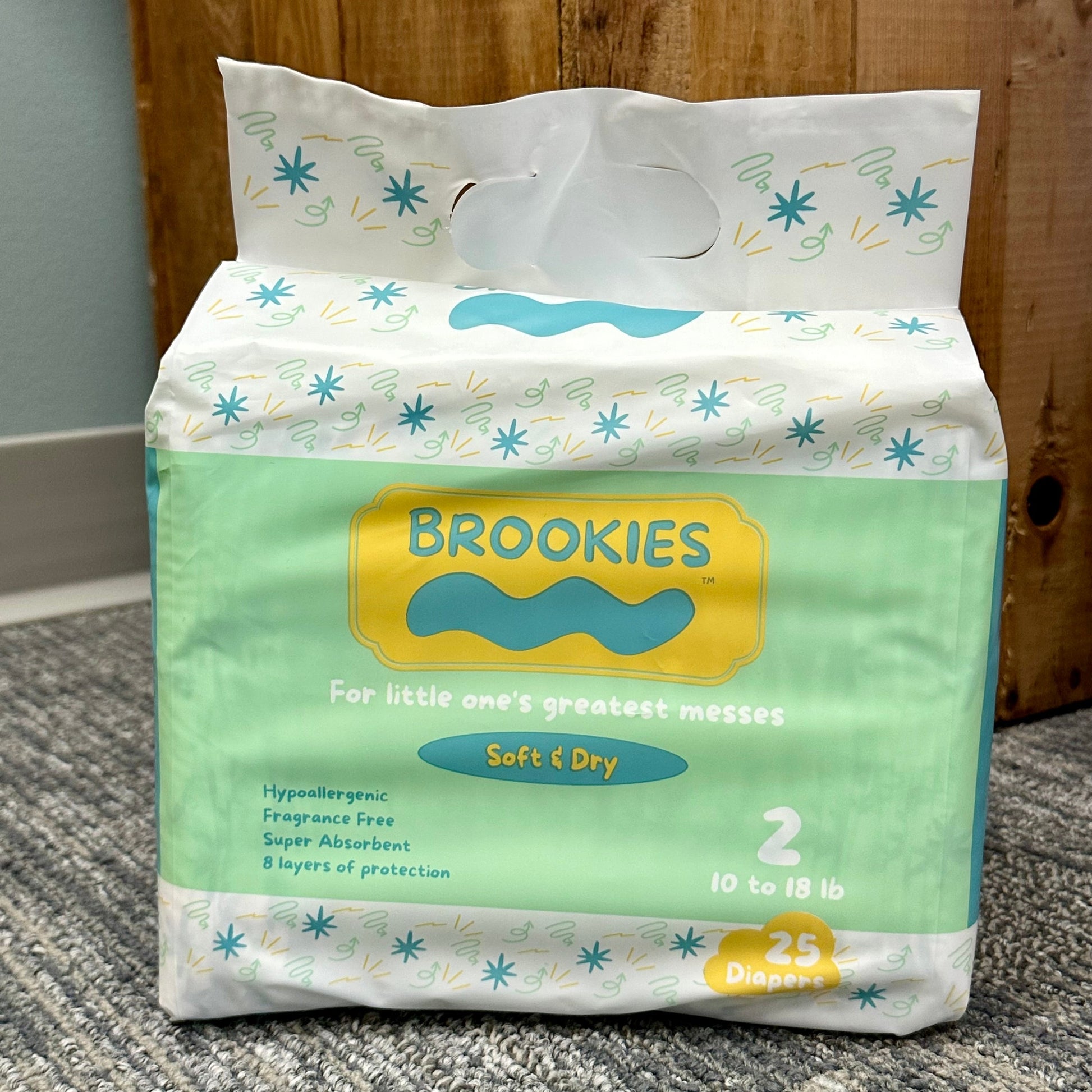 A pack of "Brookies" diapers with a yellow and green design, featuring "Soft & Dry" text, a cloud graphic, and the weight range of "10 to 18 lb," standing on a gray carpet with a wood backdrop.