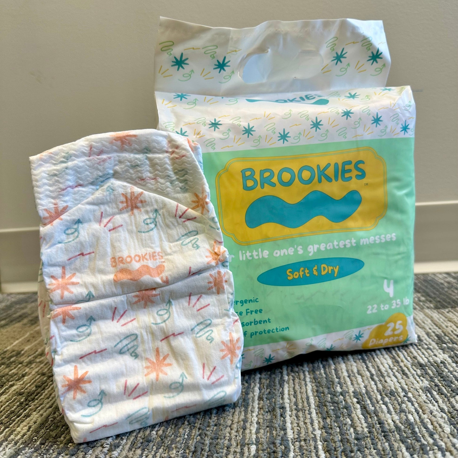 Close-up of Brookies brand diapers; a folded diaper and a pack of 25 diapers on a patterned rug.