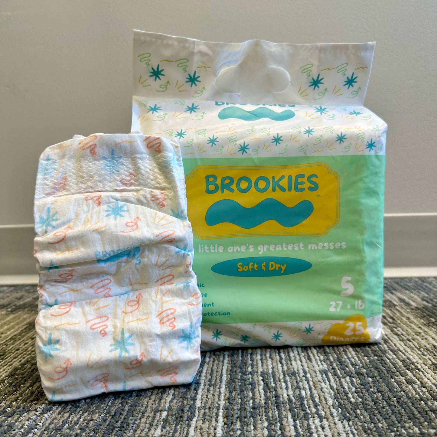 Package of Brookies size 5 diapers alongside a single folded diaper on a textured rug.