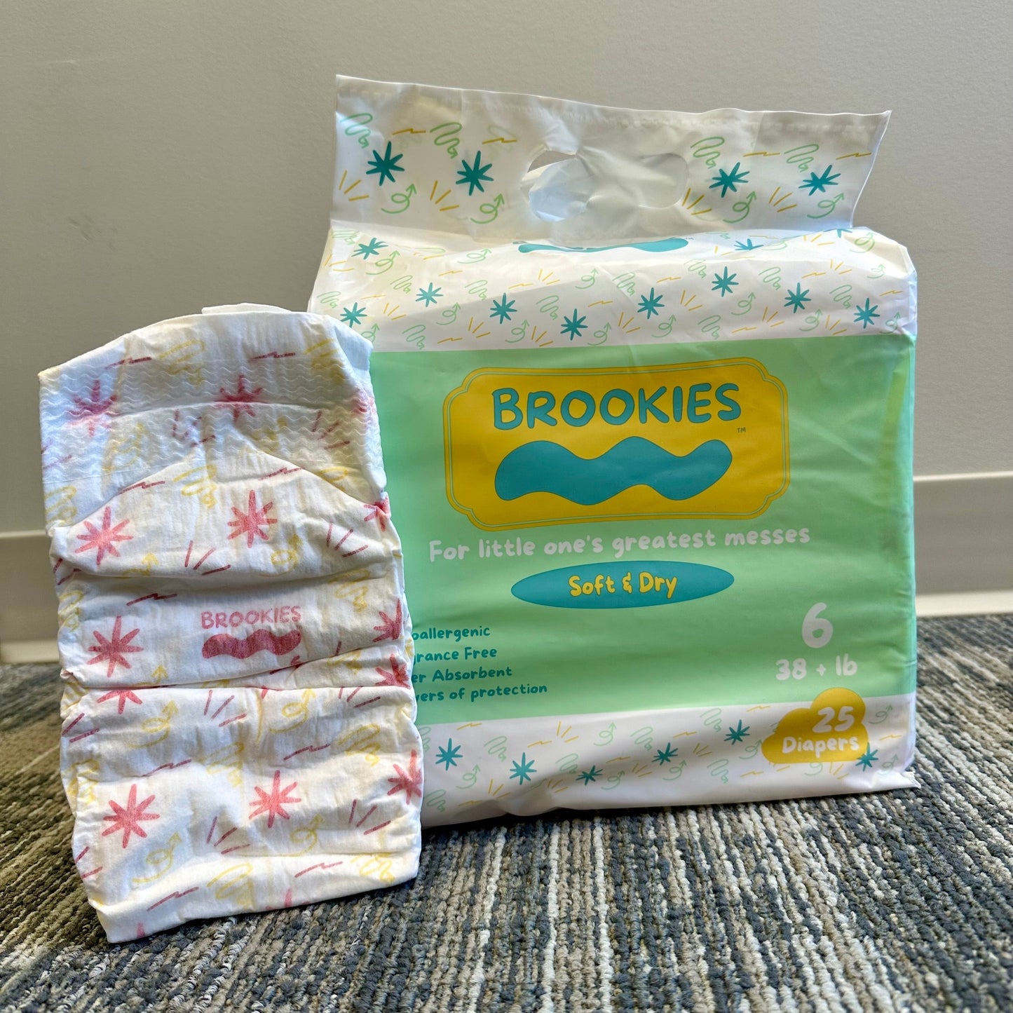 A package of Brookies brand diapers is on the right, next to a stack of diapers on the left. The bag of diapers is a light green with a yellow label, and has the words "Brookies" and "For little one's greatest messes" written on it. The stack of diapers is white with a pink and yellow design. They sit on a gray, mottled carpet.