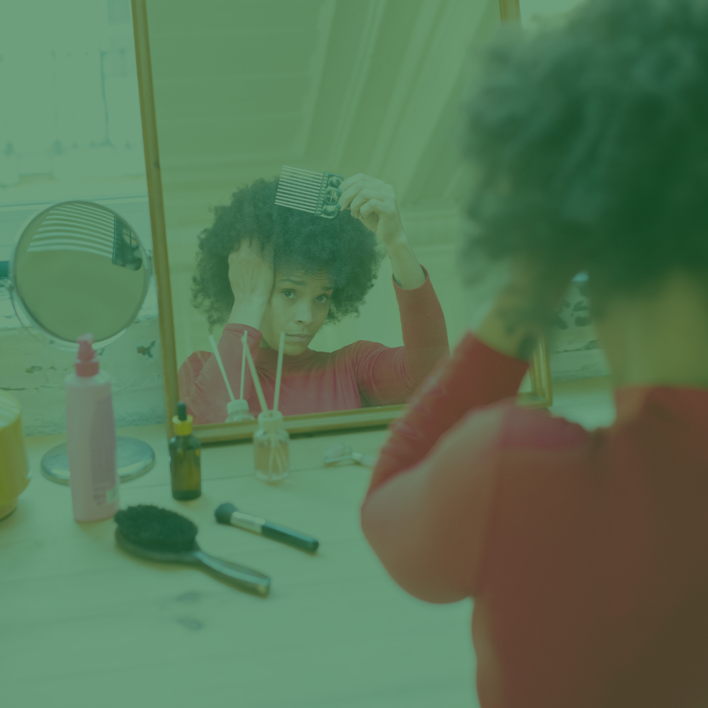 A person with curly hair looks into a bathroom mirror while combing their hair. The scene has a strong green tint and includes toiletries on the counter, with the person's reflection visible in the mirror.