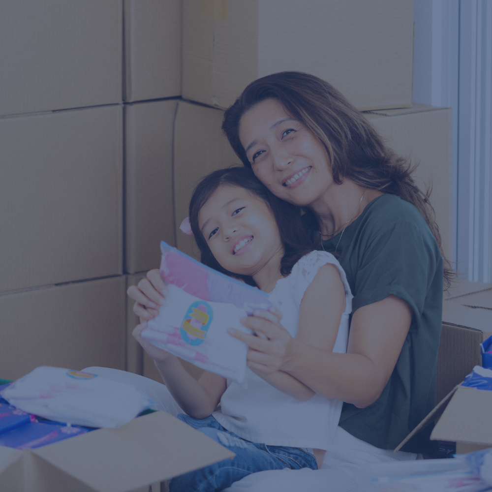 A woman and a young girl smiling while holding a pink and blue packaged item, seated in front of stacked cardboard boxes in a room.