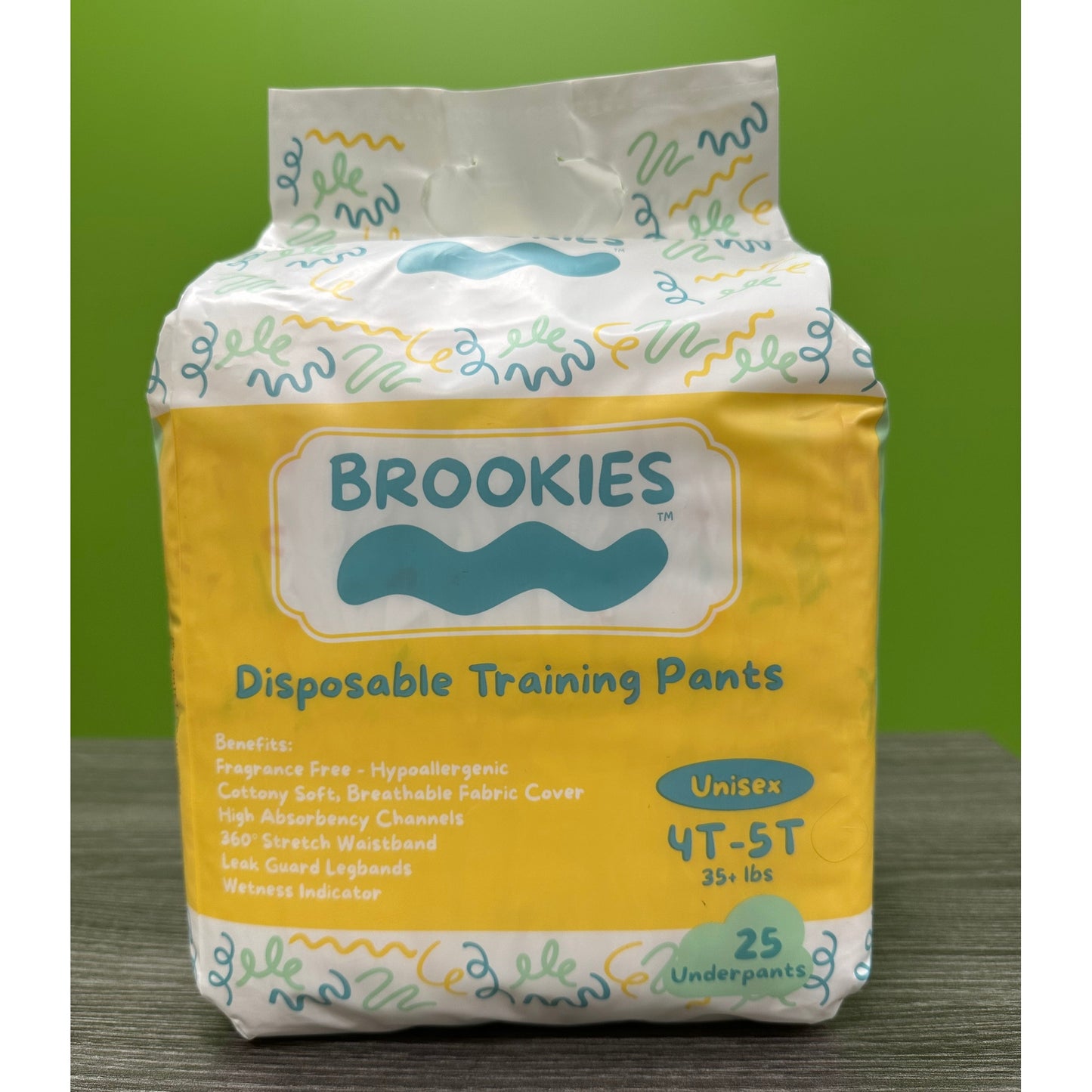 A package of Brookies disposable training pants, in yellow and white packaging with cartoonish designs, sitting on a wooden surface against a green background.