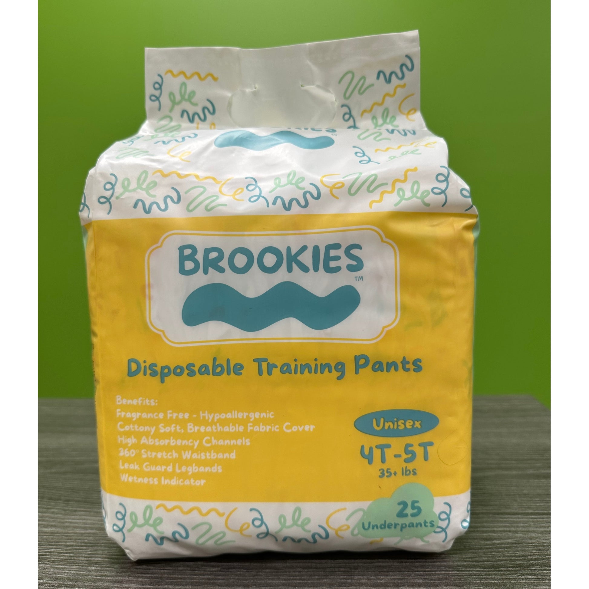 A package of Brookies disposable training pants, in yellow and white packaging with cartoonish designs, sitting on a wooden surface against a green background.