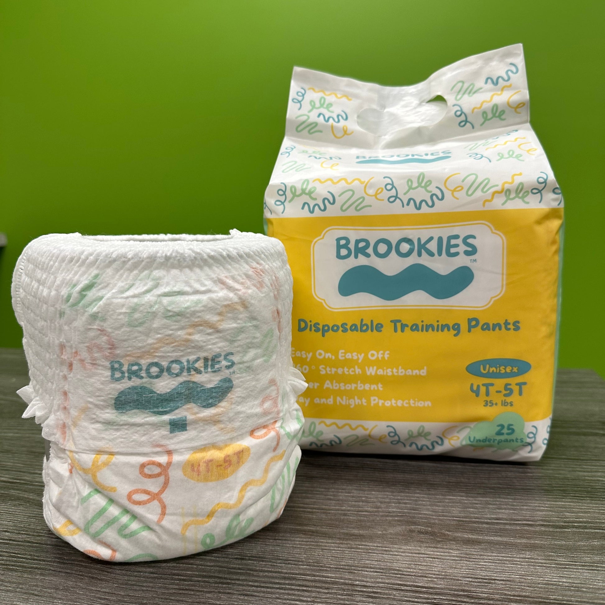 Close-up of a rolled Brookies disposable training pant in front of a closed pack of Brookies disposable training pants, set on a grey wood-like surface, with a green background.