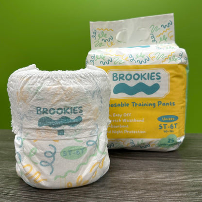 A package of Brookies training pants sits behind a single training pant on a wooden surface in front of a green background.
