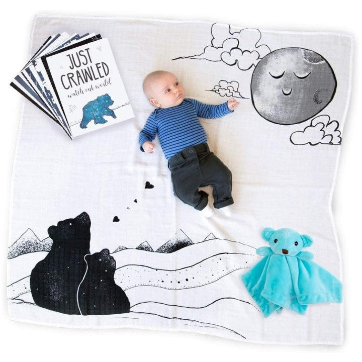 Infant lying on printed blanket with plush toy and milestone cards. The infant is wearing a blue and white striped shirt and dark grey pants. The blanket is white with an illustration of bears and a moon.