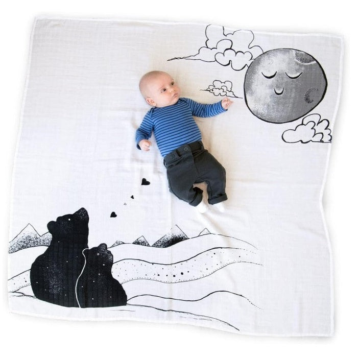 A baby in a blue striped shirt and dark pants points to a drawn moon on a white muslin blanket with black bear and mountain designs.