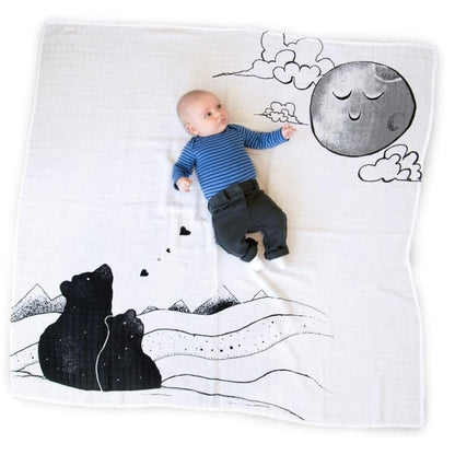 A baby in a blue striped shirt and dark pants points to a drawn moon on a white muslin blanket with black bear and mountain designs.