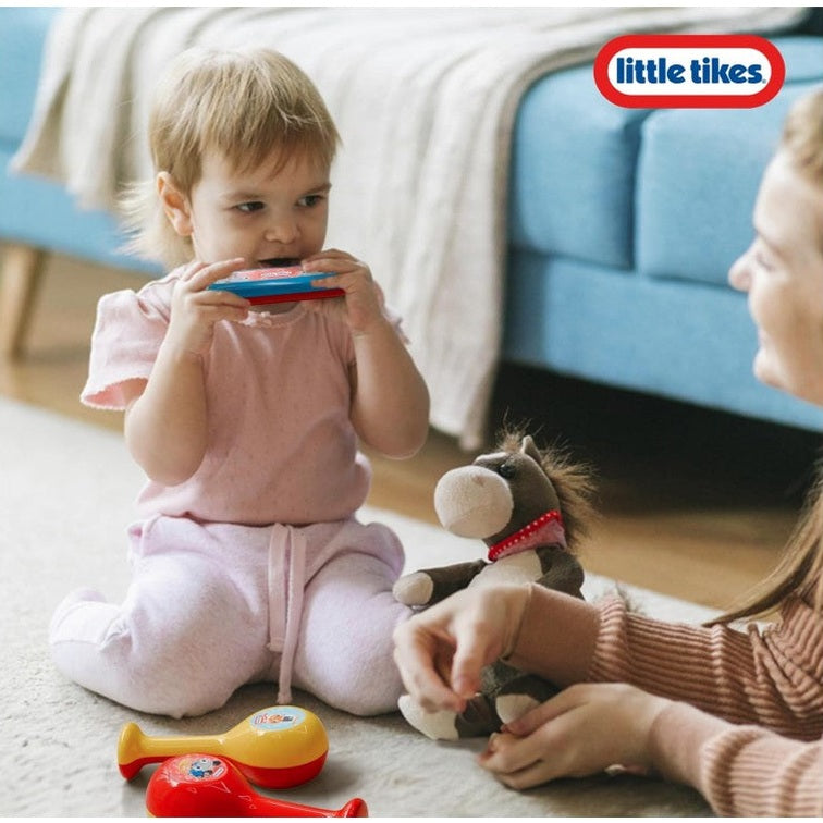 A toddler plays a harmonica while sitting on the floor next to a stuffed horse and maracas.