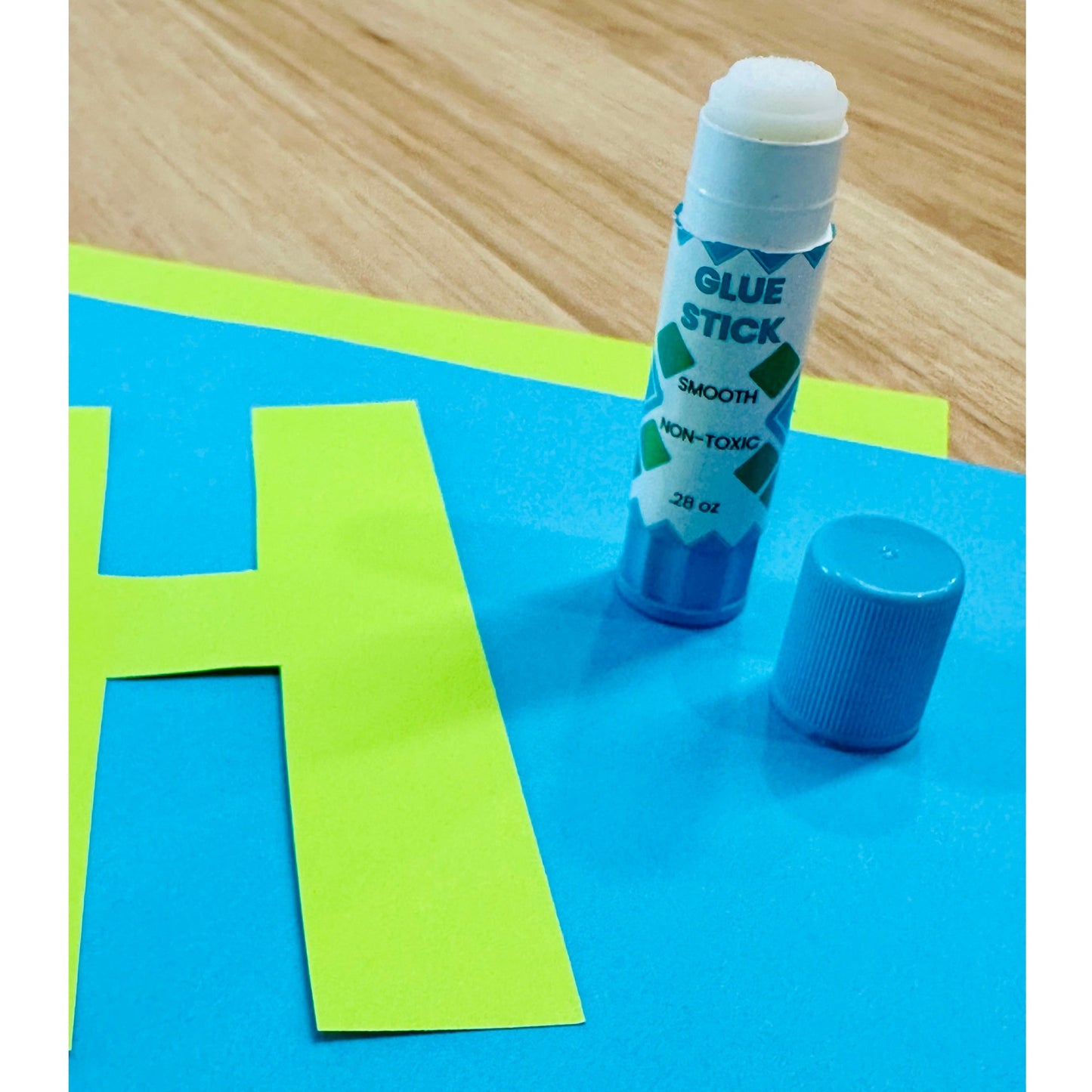 A glue stick and its cap rest on top of blue construction paper and green paper cut in the shape of a capital H.