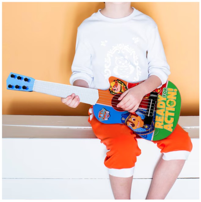 A child sitting, wearing a white shirt and orange pants, playing a Paw Patrol toy guitar.