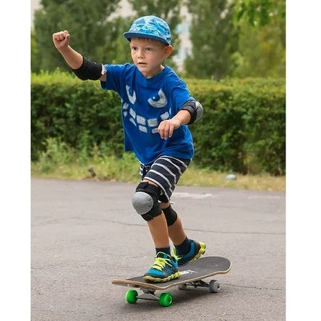 A young boy in a blue monster shirt and safety gear rides a skateboard on pavement.